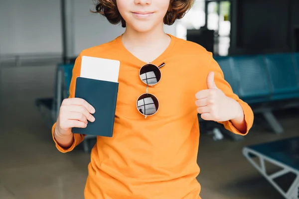 Vista cortada de menino pré-adolescente com passaporte e bilhete de ar mostrando polegar no aeroporto — Fotografia de Stock
