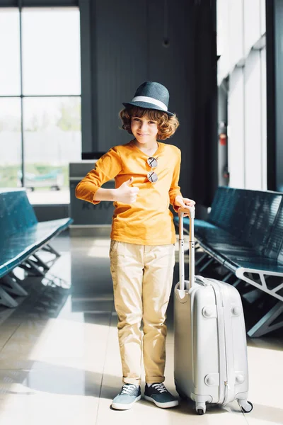 Niño preadolescente con maleta mostrando el pulgar en la sala de espera en el aeropuerto - foto de stock