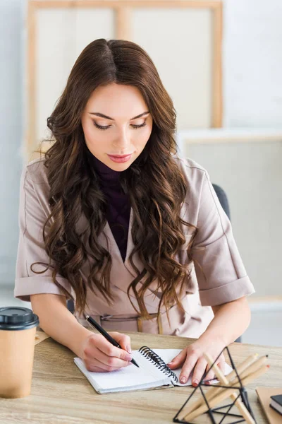 Selective focus of pretty curly girl writing in notebook near stationary and paper cup — Stock Photo
