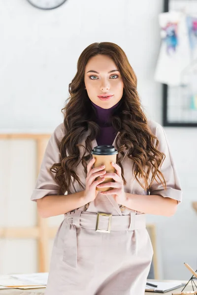 Привлекательная кудрявая молодая женщина держит бумажную чашку с кофе и смотрит в камеру — стоковое фото