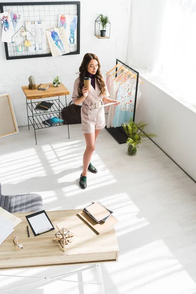 Vista aérea del diseñador rizado mirando bocetos de moda mientras sostiene la taza de papel — Stock Photo