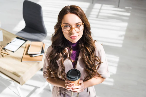 Ansicht einer glücklichen Frau mit Brille, die in die Kamera blickt, während sie Pappbecher in der Hand hält — Stockfoto