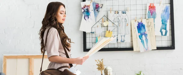 Plan panoramique de attrayant jeune designer debout avec sac près des croquis de mode — Photo de stock