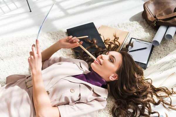 Vista aérea de la mujer pensativa acostada en la alfombra y mirando el papel cerca de bocetos de moda - foto de stock