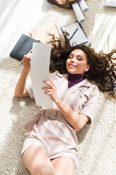 Vista superior de la mujer alegre acostada en la alfombra y mirando el papel en blanco - foto de stock