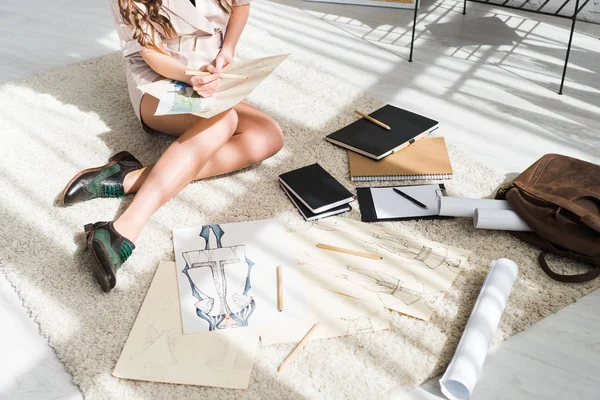 Vista cortada de jovem designer sentado no tapete e desenho perto de esboços de moda — Fotografia de Stock