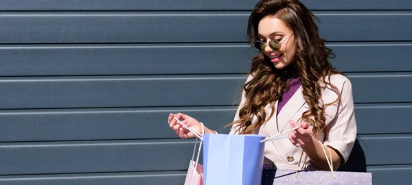 Panoramic shot of cheerful curly young woman in sunglasses looking at blue shopping bag — Stock Photo