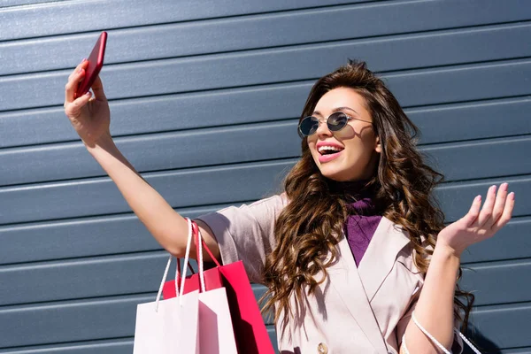 Mujer rizada feliz con bolsas de compras tomando selfie en el teléfono inteligente - foto de stock