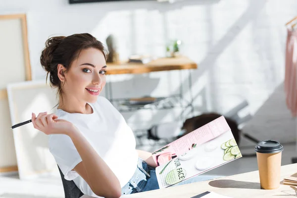 Cheerful woman in white t-shirt holding magazine and pen near paper cup on table — Stock Photo