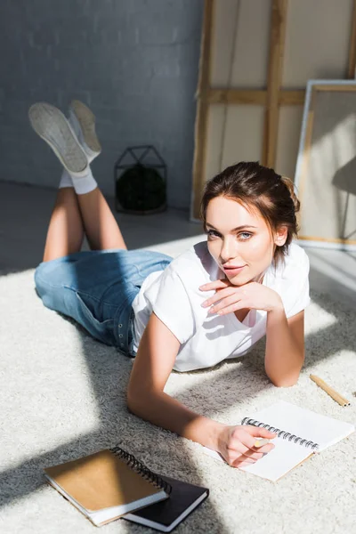 Jolie jeune femme couchée sur le tapis près des cahiers et regardant la caméra — Photo de stock