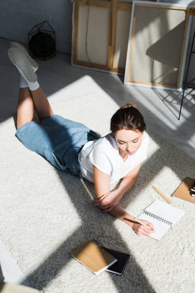 Bonita mujer joven en camiseta blanca acostada en la alfombra y la escritura en el cuaderno - foto de stock