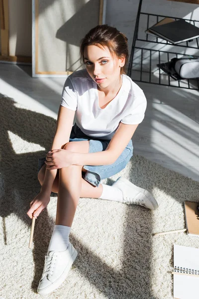 Красива молода жінка в білій футболці сидить на килимі і дивиться на камеру біля блокнотів — стокове фото
