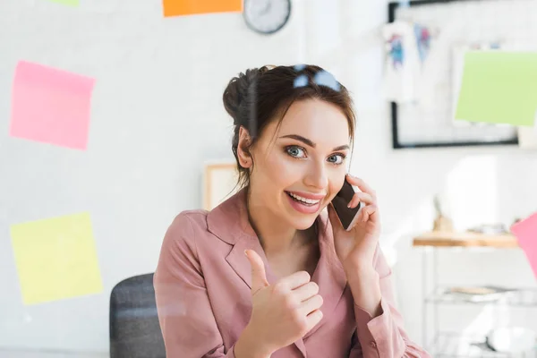 Enfoque selectivo de la mujer alegre hablando en el teléfono inteligente y mostrando el pulgar hacia arriba — Stock Photo