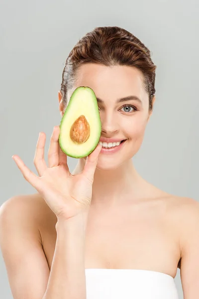 Glückliche Frau hält die Hälfte der Bio-Avocado in der Hand, während sie das Auge isoliert auf grau abdeckt — Stockfoto