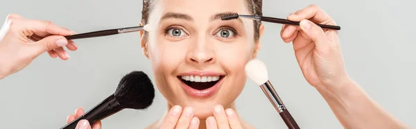 Panoramic shot of makeup artists holding cosmetic brushes near excited woman isolated on grey — Stock Photo
