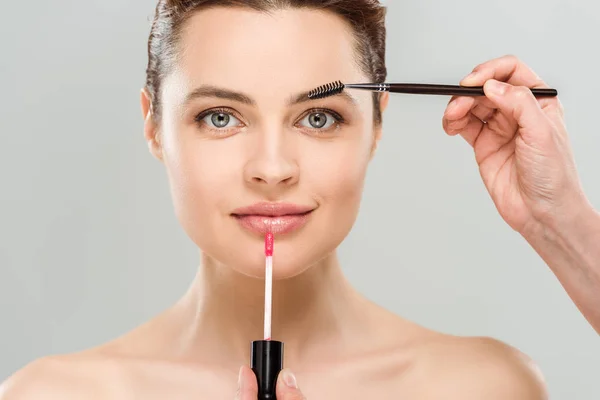 Cropped view of makeup artist holding mascara brush and lip gloss near naked woman isolated on grey — Stock Photo