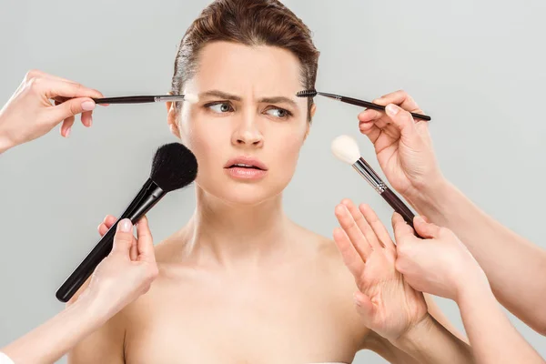 Vista cortada de maquiadores segurando escovas cosméticas perto de mulher descontente nu isolado em cinza — Fotografia de Stock