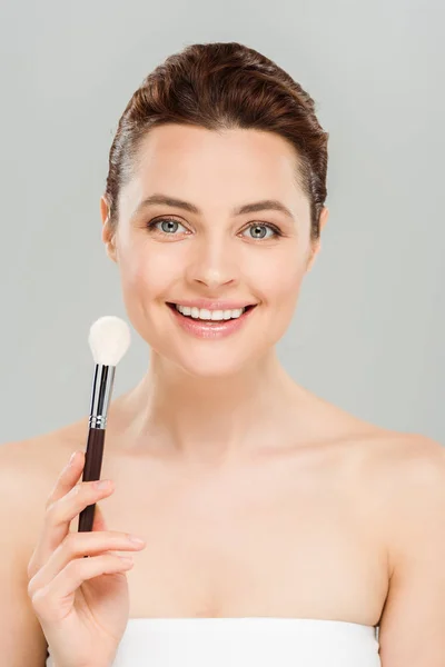 Cheerful woman holding cosmetic brush and smiling isolated on grey — Stock Photo