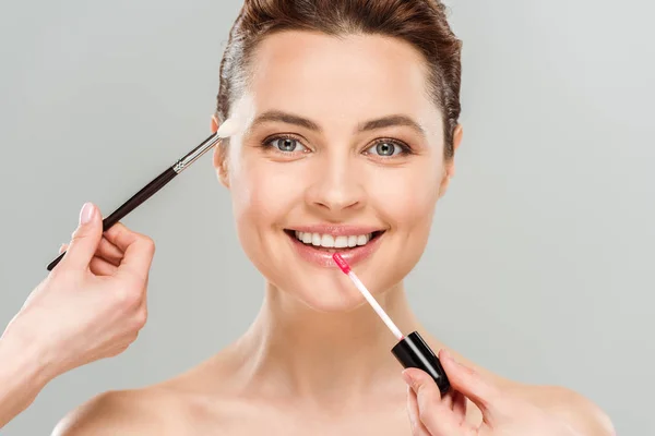 Cropped view of cosmetic artists holding lip gloss and cosmetic brush near happy naked woman isolated on grey — Stock Photo