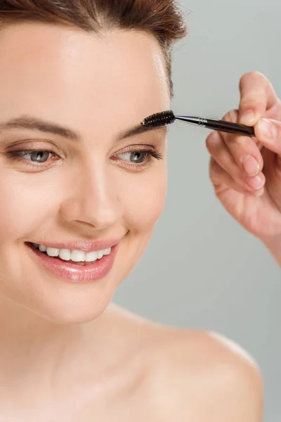 Recortado vista de artista de maquillaje sosteniendo cepillo de cejas cerca alegre mujer desnuda aislado en gris - foto de stock