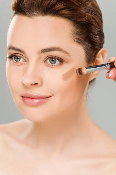 Atractiva mujer sosteniendo cepillo cosmético y aplicando rubor marrón en la mejilla aislado en gris - foto de stock