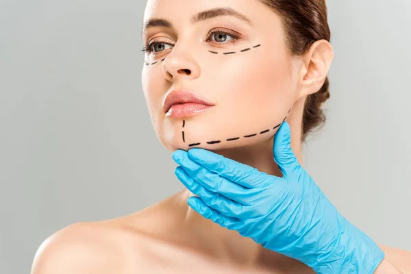Vista recortada del cirujano plástico tocando la cara de la mujer atractiva con la cara marcada aislada en gris - foto de stock