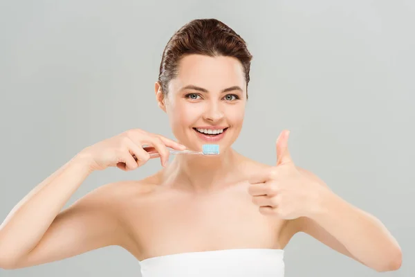 Mujer alegre mostrando el pulgar hacia arriba mientras sostiene el cepillo de dientes aislado en gris - foto de stock