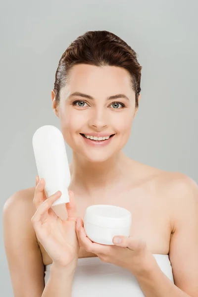 Alegre mujer sosteniendo contenedor con crema cosmética y botella de loción corporal aislado en gris - foto de stock