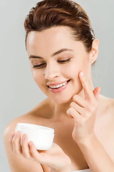 Donna felice guardando contenitore durante l'applicazione di crema viso isolato su grigio — Foto stock