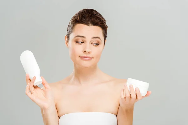 Atractiva mujer mirando contenedor con crema cosmética y la celebración de la botella de loción corporal aislado en gris - foto de stock