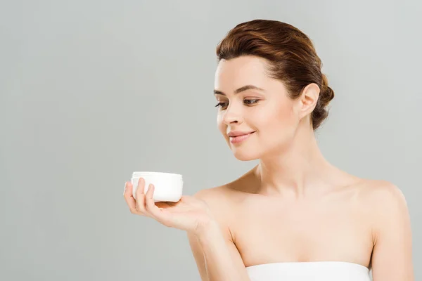 Cheerful woman looking at container with cosmetic cream isolated on grey — Stock Photo