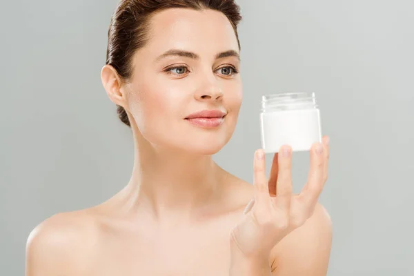 Cheerful naked woman looking at container with cosmetic cream isolated on grey — Stock Photo