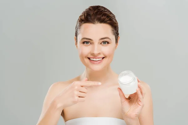 Mujer feliz señalando con el dedo al contenedor con crema facial y mirando a la cámara aislada en gris - foto de stock