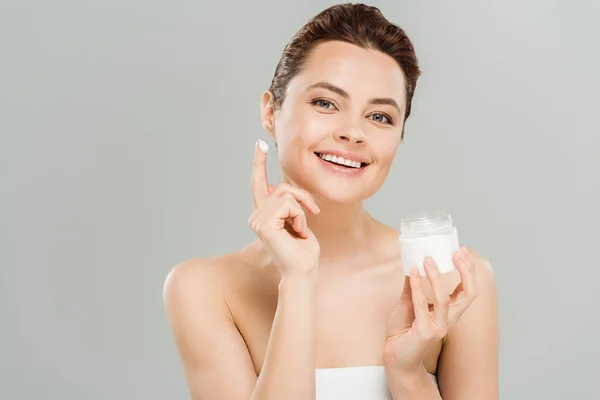 Mujer feliz sosteniendo contenedor con crema facial y mirando a la cámara aislada en gris - foto de stock