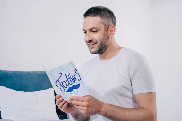 Feliz hombre sosteniendo padres día tarjeta de felicitación hecha a mano mientras está sentado en el dormitorio - foto de stock