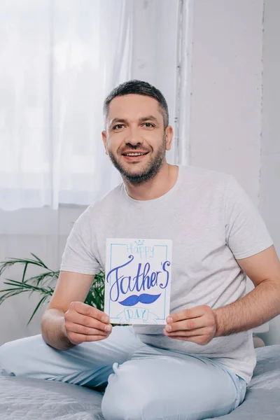 Glücklicher Mann zeigt Vatertags-Grußkarte, während er zu Hause im Bett sitzt — Stockfoto