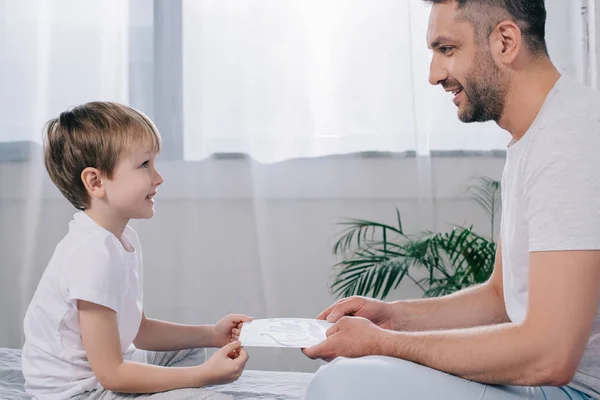 Seitenansicht des netten Jungen, der dem glücklichen Papa eine Grußkarte zum Vatertag schenkt — Stockfoto