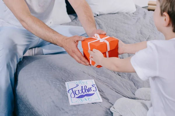 Abgeschnittene Aufnahme eines Kindes, das seinem auf Bettwäsche sitzenden Vater eine Geschenkbox zum Vatertag überreicht — Stockfoto