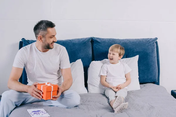 Happy man holding fathers day gift box while sitting on bed near cheerful son — Stock Photo