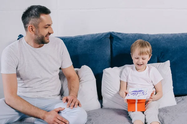 Entzückender Junge mit Geschenkbox und Grußkarte zum Vatertag, während er auf dem Bett in der Nähe eines glücklichen Vaters sitzt — Stockfoto