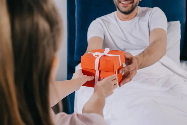 Vista parcial del hombre sonriente que toma la caja de regalo del día de los padres de la hija mientras está sentado debajo de la manta - foto de stock