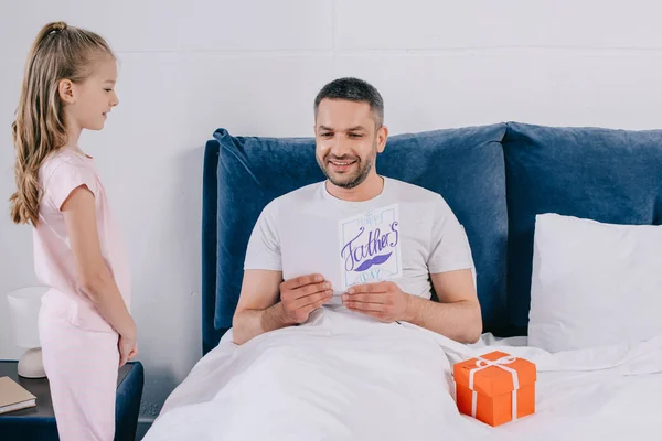 Cute child standing near father holding fathers day greeting card while lying in bed — Stock Photo