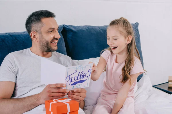 Felice uomo in possesso di scatola regalo e padri giorno biglietto di auguri mentre seduto vicino a figlia allegra — Foto stock