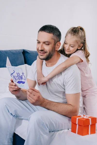 Lindo niño abrazando feliz papá sosteniendo padres día tarjeta de felicitación mientras está sentado cerca de la caja de regalo - foto de stock