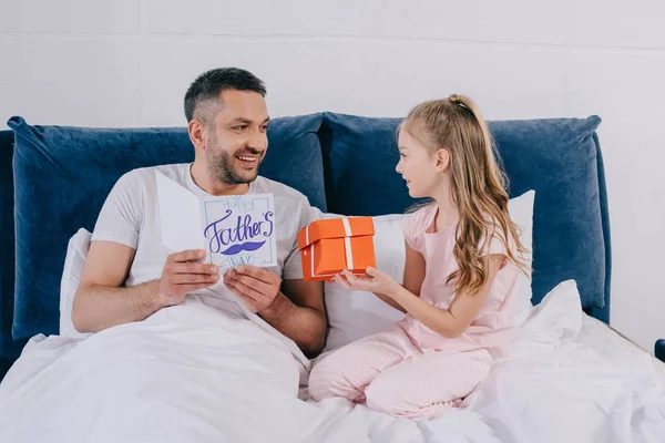 Feliz hombre sosteniendo padres día tarjeta de felicitación mientras está sentado cerca de la hija sosteniendo caja de regalo - foto de stock