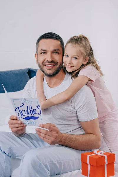 Entzückende Tochter umarmt glücklichen Vater mit Vatertags-Grußkarte, während sie in der Nähe von Geschenkbox sitzt — Stockfoto