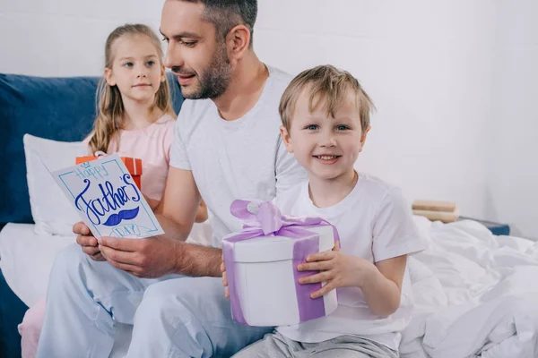 Прелестная дочь и сын сидят с подарочными коробками рядом с счастливым папочкой держа открытку на день отца — стоковое фото