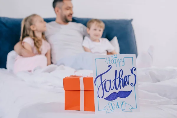 Selective focus of red gift box and happy fathers day greeting card near cheerful man embracing son and daughter while sitting on bed together — Stock Photo