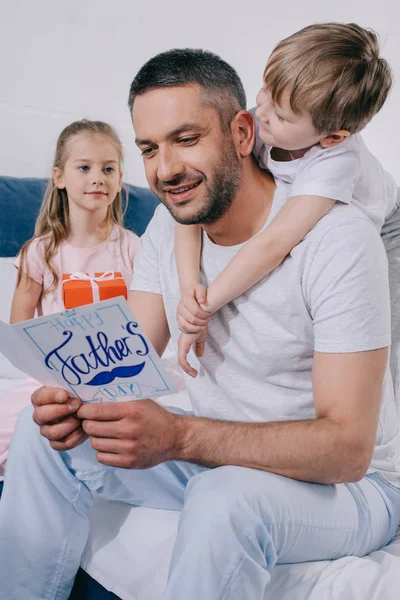 Menino bonito abraçando o pai olhando para o dia dos pais cartão perto adorável filha com caixa de presente — Fotografia de Stock
