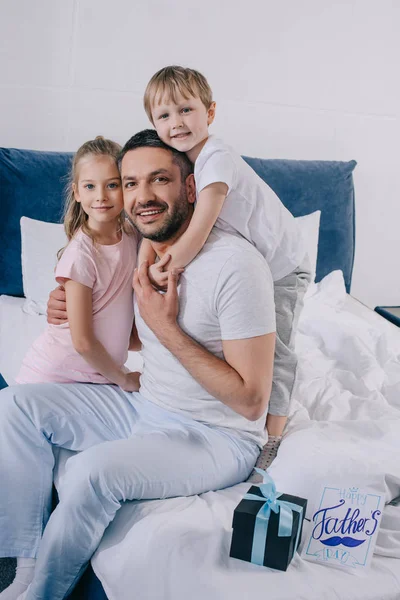 Adorables niños abrazando feliz padre cerca de padres día tarjeta de felicitación y caja de regalo en la ropa de cama - foto de stock